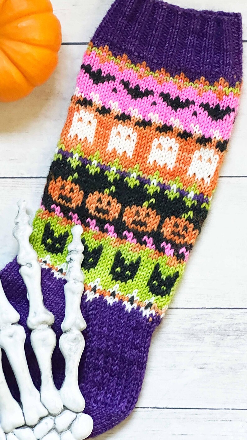 Puede incluir: Un calcet&iacute;n de Halloween tejido de color p&uacute;rpura y multicolor con murci&eacute;lagos, fantasmas, calabazas y gatos. El calcet&iacute;n presenta un pu&ntilde;o acanalado y un dise&ntilde;o estampado en rosa, naranja, negro y verde. Una mano de esqueleto decorativa est&aacute; en primer plano.