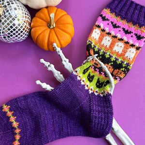 Puede incluir: Un par de calcetines morados de punto con dise&ntilde;os de Halloween, incluyendo fantasmas y murci&eacute;lagos, exhibidos con una mano de esqueleto, calabazas y una bola de discoteca sobre un fondo morado. Los calcetines tienen detalles en naranja, verde y negro.