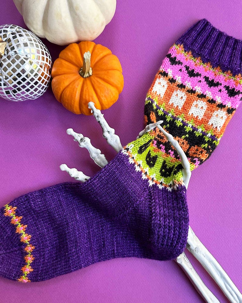 Puede incluir: Un par de calcetines morados de punto con dise&ntilde;os de Halloween, incluyendo fantasmas y murci&eacute;lagos, exhibidos con una mano de esqueleto, calabazas y una bola de discoteca sobre un fondo morado. Los calcetines tienen detalles en naranja, verde y negro.