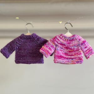 Two miniature hand-knitted sweater ornaments hanging on tiny wooden hangers. One sweater is a deep purple, the other is a multicolored pink, purple, and white yarn.  Both are long-sleeved crew neck sweaters.