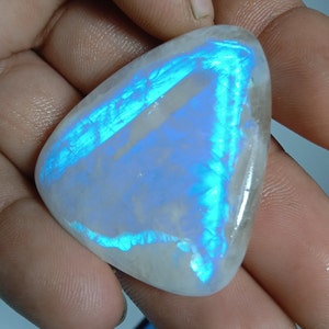 Puede incluir: Una gema pulida, de forma triangular, con una base blanca nacarada y destellos iridiscentes azules y turquesas. La piedra se sostiene en una mano, mostrando su superficie lisa y reflectante. Se destaca el juego de colores único de la gema.