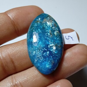 Puede incluir: Gema ovalada pulida, de un vibrante color azul con inclusiones brillantes. La piedra se sostiene en una mano, mostrando su superficie lisa y belleza natural. Una pequeña etiqueta blanca con el número "5" es visible.