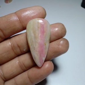 Peut inclure: Une pierre précieuse polie en forme de goutte d'eau. La pierre présente un mélange de couleurs blanc crème et rose, avec une bande rose prononcée au centre. Le bijou est tenu dans une main, mettant en valeur sa forme et sa taille.