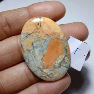 Puede incluir: Una gema ovalada con un patrón único de tonos naranja, grises y amarillos. La piedra tiene una apariencia agrietada, con una mezcla de colores y texturas. La gema se sostiene en una mano.