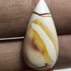 Puede incluir: Una gema pulida en forma de lágrima. Presenta una mezcla de colores: blanco, amarillo y marrón rojizo, con una punta blanca. El diseño tiene un patrón de rayas. Esta gema se utiliza probablemente para joyería.
