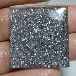 Puede incluir: Un elemento decorativo cuadrado gris y plateado con purpurina. La superficie está llena de partículas brillantes de purpurina plateada y gris, creando un efecto brillante. Los bordes son lisos y pulidos.