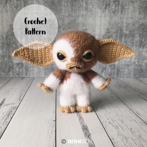 May include: A handmade crochet creature with large ears, big black eyes, and a mischievous expression. The plush toy is brown, white, and tan, with the words "Crochet Pattern" in a white circle. The toy is standing on a white wooden surface.