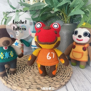 Puede incluir: Juguetes de animales amigurumi de crochet, incluyendo una rana roja con una camiseta amarilla y el número 67, un oso marrón con un suéter verde, un conejo blanco con una camiseta azul y una estrella, y un perezoso marrón con una camiseta amarilla. Los juguetes están sentados en una estera de paja tejida.