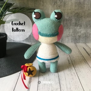 Puede incluir: Patrón de crochet para una rana verde que lleva un vestido blanco con ribetes azules. La rana tiene mejillas rosadas y ojos marrones. Una pequeña bolsa de monedas de oro con una cinta roja está en primer plano.