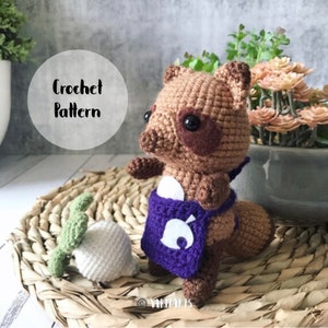 Puede incluir: Juguete de peluche de mapache tejido a crochet con un delantal morado con un diseño de hoja blanca. El mapache es marrón con ojos negros y una nariz marrón. El juguete está sentado en una superficie de mimbre tejido con un nabo blanco tejido a crochet.