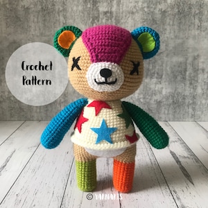 Puede incluir: Juguete de peluche de crochet de un oso con pelo rosa, pelaje marrón, ropa azul, verde y blanca, y una estrella blanca en el pecho. El juguete está de pie sobre una superficie blanca. El texto "Crochet Pattern" es visible en el fondo.