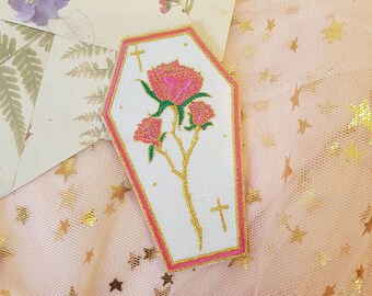 Könnte beinhalten: Ein weißer, sargförmiger Patch mit einer rosa Rose und einer goldenen Kreuzstickerei. Der Patch hat einen rosa Rand und befindet sich auf einem rosa Hintergrund mit goldenen Sternen.