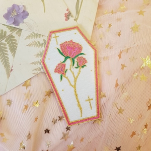 Könnte beinhalten: Ein weißer, sargförmiger Patch mit einer rosa Rose und einer goldenen Kreuzstickerei. Der Patch hat einen rosa Rand und befindet sich auf einem rosa Hintergrund mit goldenen Sternen.