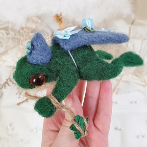 May include: A green felt frog wearing a blue felt hat with a blue and white ribbon. The frog has a brown eye and is holding a green and brown felt leaf in its hand. The frog is flying with a blue felt cape.