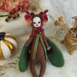 May include: A handmade felt doll with brown fur, green wings, and a red and green ribbon. The doll has a painted face with red cheeks and a flower crown with red and pink flowers.