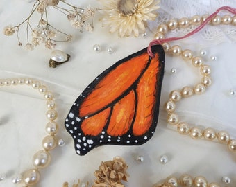 May include: A hand-painted wooden butterfly ornament with orange and black wings. The ornament is attached to a string of pearls and a pink ribbon.