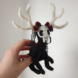 Può includere: Un animale di feltro nero con una testa di teschio bianca e corna. L'animale indossa una corona di fiori bordeaux e un velo di pizzo nero.