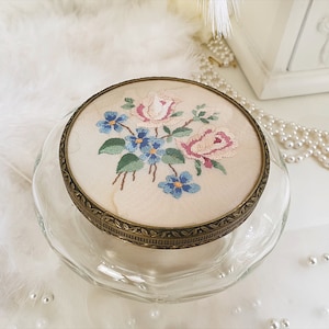 Peut inclure: Une boîte à bijoux vintage en verre et en laiton avec un motif floral. Le couvercle est décoré de fleurs brodées roses et bleues sur un fond crème.