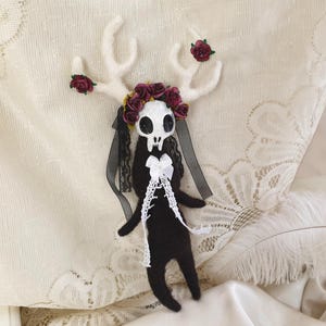 May include: A black felt doll with a white skull head and antlers. The doll is wearing a black dress with white lace and a wreath of burgundy flowers.