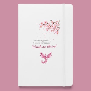Puede incluir: Diario blanco con un diseño floral rosa y el texto "I survived my period. I'll survive menopause. Watch me thrive!" También presenta una ilustración de un fénix rosa. El diario tiene un cierre de banda elástica blanca.