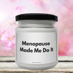 Könnte beinhalten: Ein weißes Kerzengefäß mit schwarzem Deckel und einem weißen Etikett mit der Aufschrift "Menopause Made Me Do It" und dem Text "@MenopauseCoach" darunter.