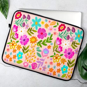 May include: A laptop sleeve with a vibrant floral pattern. The design features a light pink background adorned with colorful flowers in shades of pink, orange, blue, and purple, with green leaves. The sleeve has a black zipper and trim.