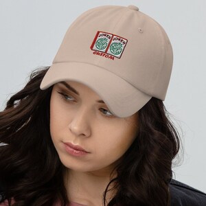 Peut inclure: Une casquette de baseball beige avec un motif brodé de deux boîtes rouges avec le mot "JOKER" et le mot "CUSTOM" en dessous.