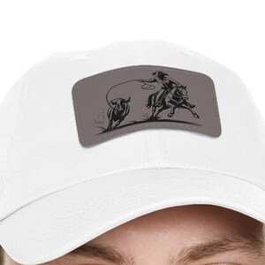 May include: White baseball cap with a gray patch featuring a black illustration of a cowboy on horseback roping a bull. The cowboy is wearing a hat and the bull is running.