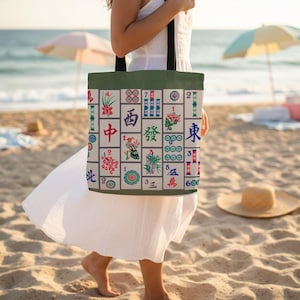 Puede incluir: Un bolso tote beige con un borde superior verde que presenta un colorido patrón de fichas de Mahjong. El bolso tiene asas negras y lo lleva una persona en una playa de arena con el océano al fondo.