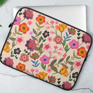 May include: A floral laptop sleeve featuring a beige background with a colorful watercolor design. The pattern includes orange, pink, purple, and yellow flowers with green leaves. The sleeve has a black zipper and trim.