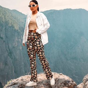 May include: A woman wearing a white denim jacket and black pants with a floral pattern. She is standing on a rocky cliff with a mountain range in the background.