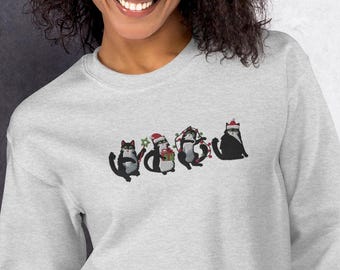 May include: A light grey sweatshirt with a design of four black cats in festive Christmas attire. The cats are wearing Santa hats and fairy lights, creating a cheerful holiday design, perfect for the festive season.