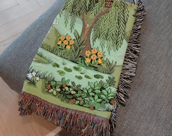 May include: A woven throw blanket featuring a nature scene with a willow tree, flowers, and a pond. The blanket has a green background with a colorful fringe border. The design includes a variety of plants and a small owl.