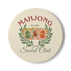 Pode incluir: Uma base para copos redonda, esbranquiçada, com as palavras "MAHJONG Social Club" em letras vermelhas e verdes. A base para copos apresenta três peças de Mahjong ilustradas e o texto "EST 2026". Uma grinalda verde envolve as peças.
