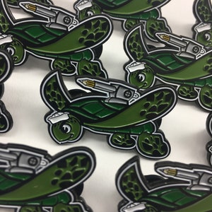 May include: A collection of green and black enamel pins featuring a cartoon turtle wearing a sailor hat and holding a missile. The turtle is on a green and black shell with a black web pattern.