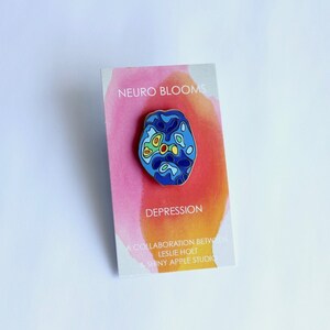 Op de afbeelding: Een blauwe en gele emaille pin met een gestileerd brein met de tekst "NEURO BLOOMS DEPRESSION" en "A COLLABORATION BETWEEN LESLIE HOLT & SHINY APPLE STUDIO" op een roze en oranje achtergrond.