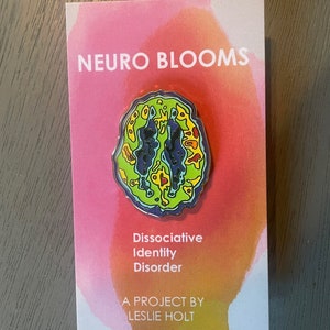 Op de afbeelding: Een kleurrijke emaille pin met een afbeelding van een hersenen met de tekst "Neuro Blooms Dissociative Identity Disorder A Project by Leslie Holt" op een roze en oranje achtergrond.