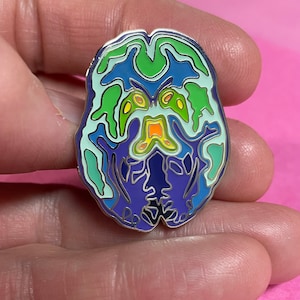 May include: A silver enamel pin shaped like a brain with a colourful, detailed anatomical design. The brain is outlined in silver and filled with shades of blue, green, yellow, orange, and purple.