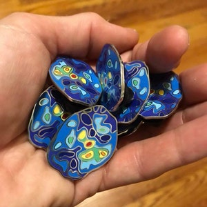 Op de afbeelding: Een verzameling blauwe, gele, groene en rode emaille-pins met een wervelend, abstract ontwerp. De pins zijn rond en hebben een zilveren metalen achterkant.