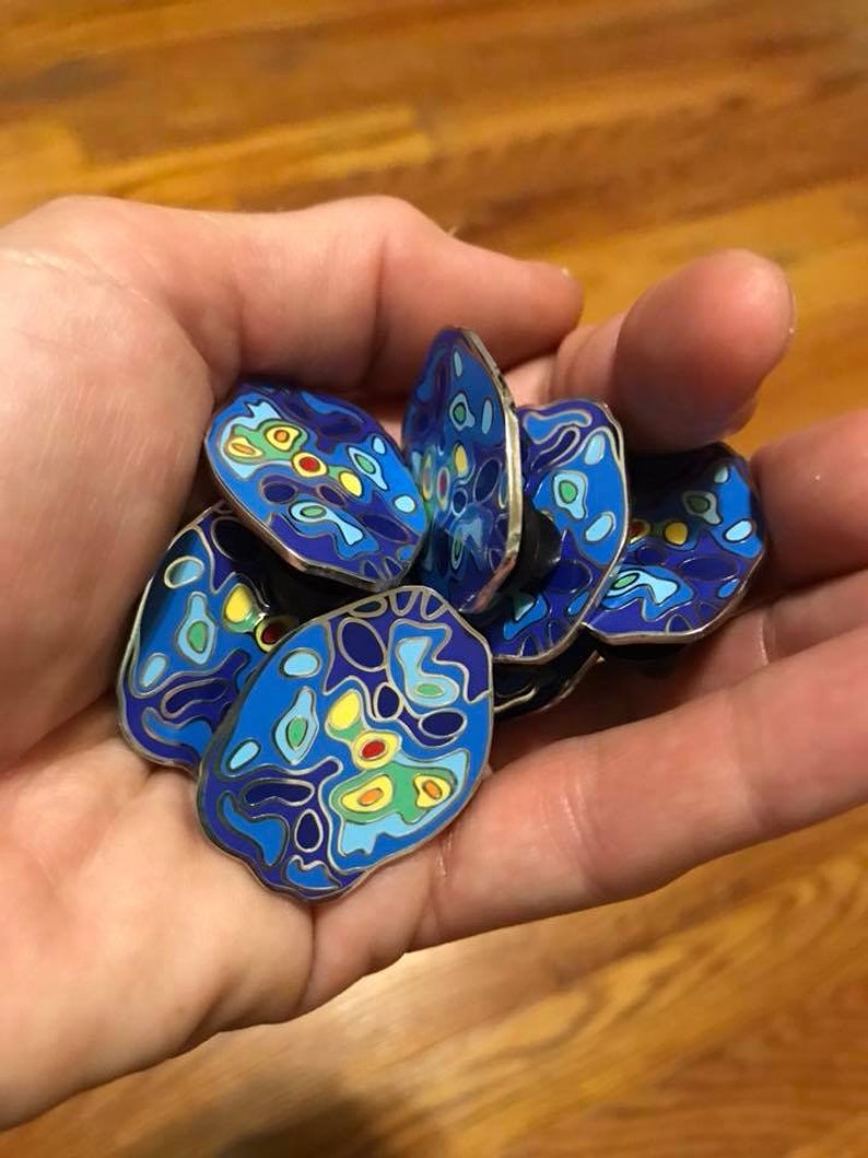 Op de afbeelding: Een verzameling blauwe, gele, groene en rode emaille-pins met een wervelend, abstract ontwerp. De pins zijn rond en hebben een zilveren metalen achterkant.