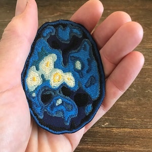 Pode incluir: Um remendo bordado azul e branco com um design em espiral. O remendo tem formato oval e tem um contorno preto.