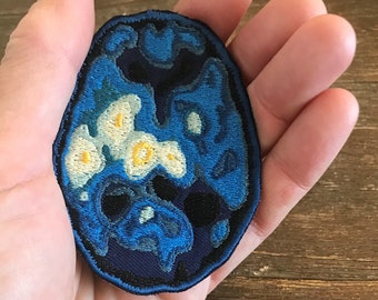 May include: A blue and white embroidered patch with a swirling design. The patch is shaped like an oval and has a black outline.