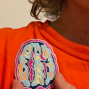 May include: A colorful embroidered patch of a brain with a white outline. The patch is on a bright orange shirt.