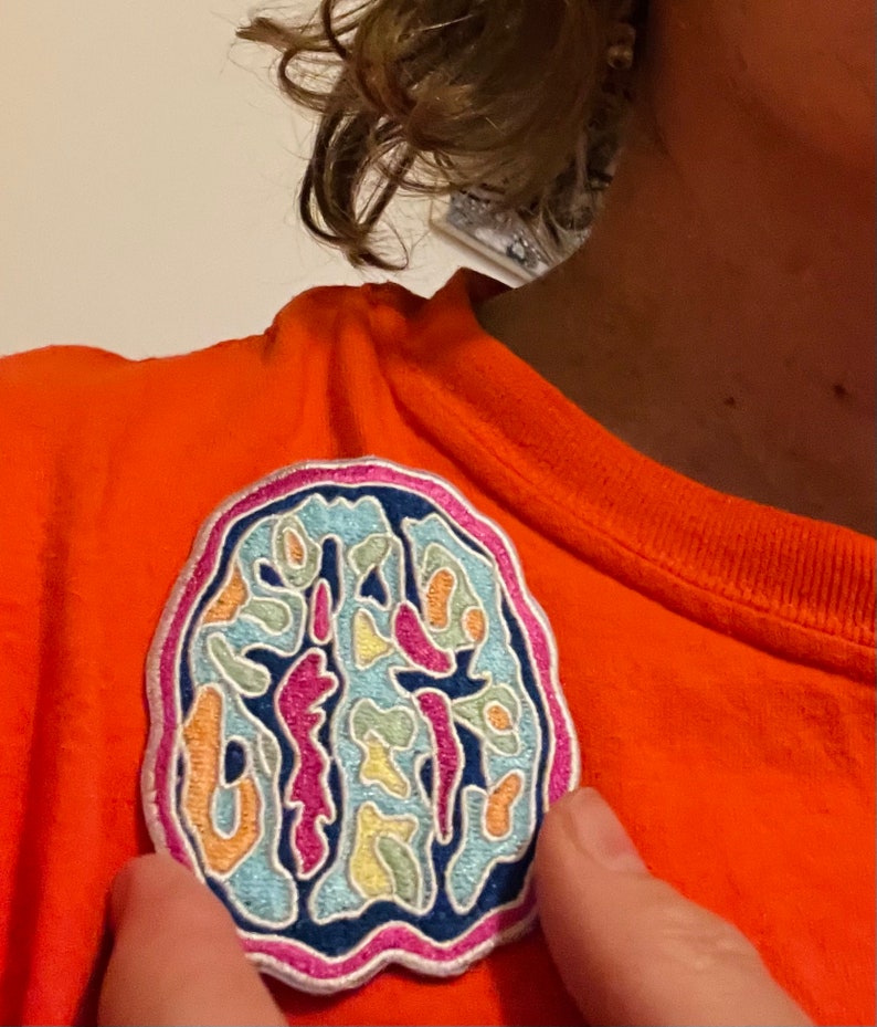 May include: A colorful embroidered patch of a brain with a white outline. The patch is on a bright orange shirt.