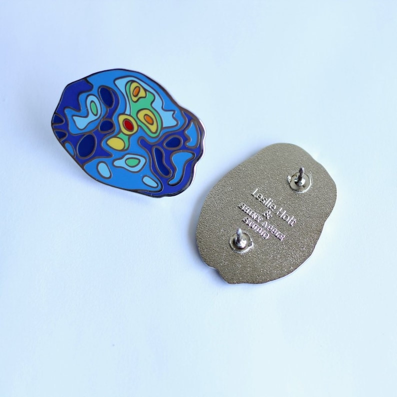 Op de afbeelding: Een blauwe en gele emaille pin met een topografisch kaartontwerp. De achterkant van de pin is zilver en heeft de tekst "Leslie Holt & Steve Aegers Studio" gegraveerd.