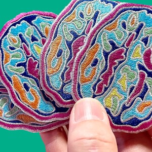 May include: Four colorful embroidered patches with a swirling, abstract design in shades of blue, pink, orange, yellow, and green. The patches are layered on top of each other and have a pink, white, and blue border.