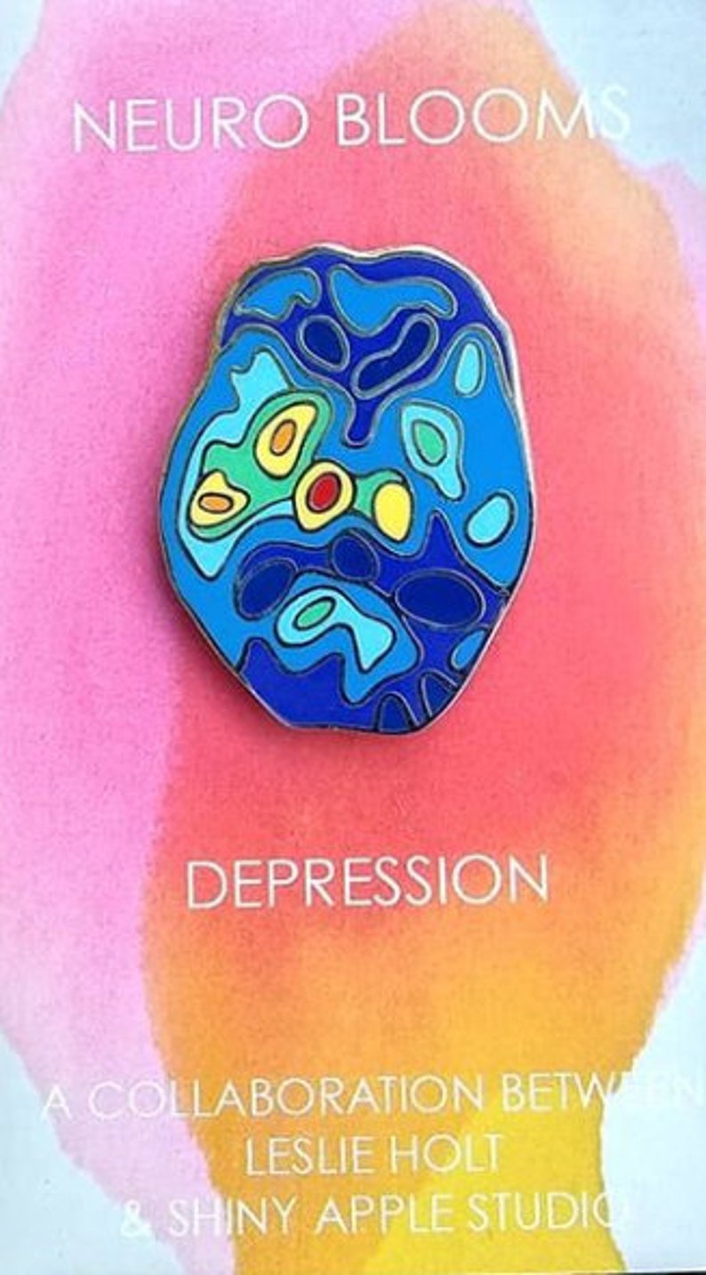 Op de afbeelding: Een blauwe en gele emaille pin met een gestileerd brein met de tekst "DEPRESSION" en "NEURO BLOOMS" eronder. De pin is een samenwerking tussen Leslie Holt en Shiny Apple Studio.