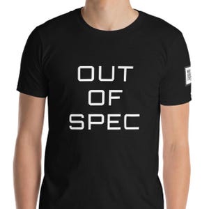 May include: A black short-sleeved t-shirt featuring the white text OUT OF SPEC centered on the chest in a bold, sans-serif font. A small square logo patch with white text is visible on the left sleeve near the shoulder seam.