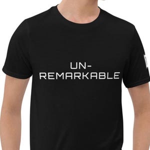 May include: A black short-sleeved t-shirt featuring the white text UN-REMARKABLE printed in a clean, sans-serif font across the chest. The garment has a classic crew neckline and a small white rectangular label on the left sleeve.