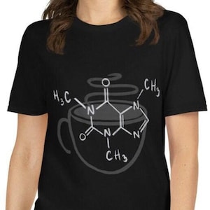 May include: A black short-sleeved t-shirt featuring a white graphic print of a coffee cup combined with the chemical structure of caffeine, including the molecular formula components H3C and CH3 written in white ink across the center chest area.